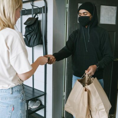 A masked courier hands over shopping bags to a customer in a contactless delivery exchange.