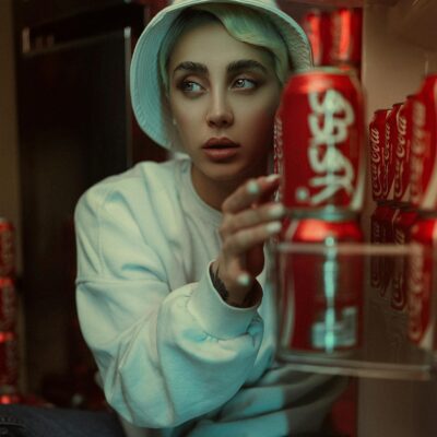 Stylish model with hat and pullover sitting near Coca-Cola cans indoors.