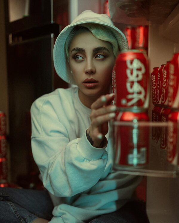Stylish model with hat and pullover sitting near Coca-Cola cans indoors.