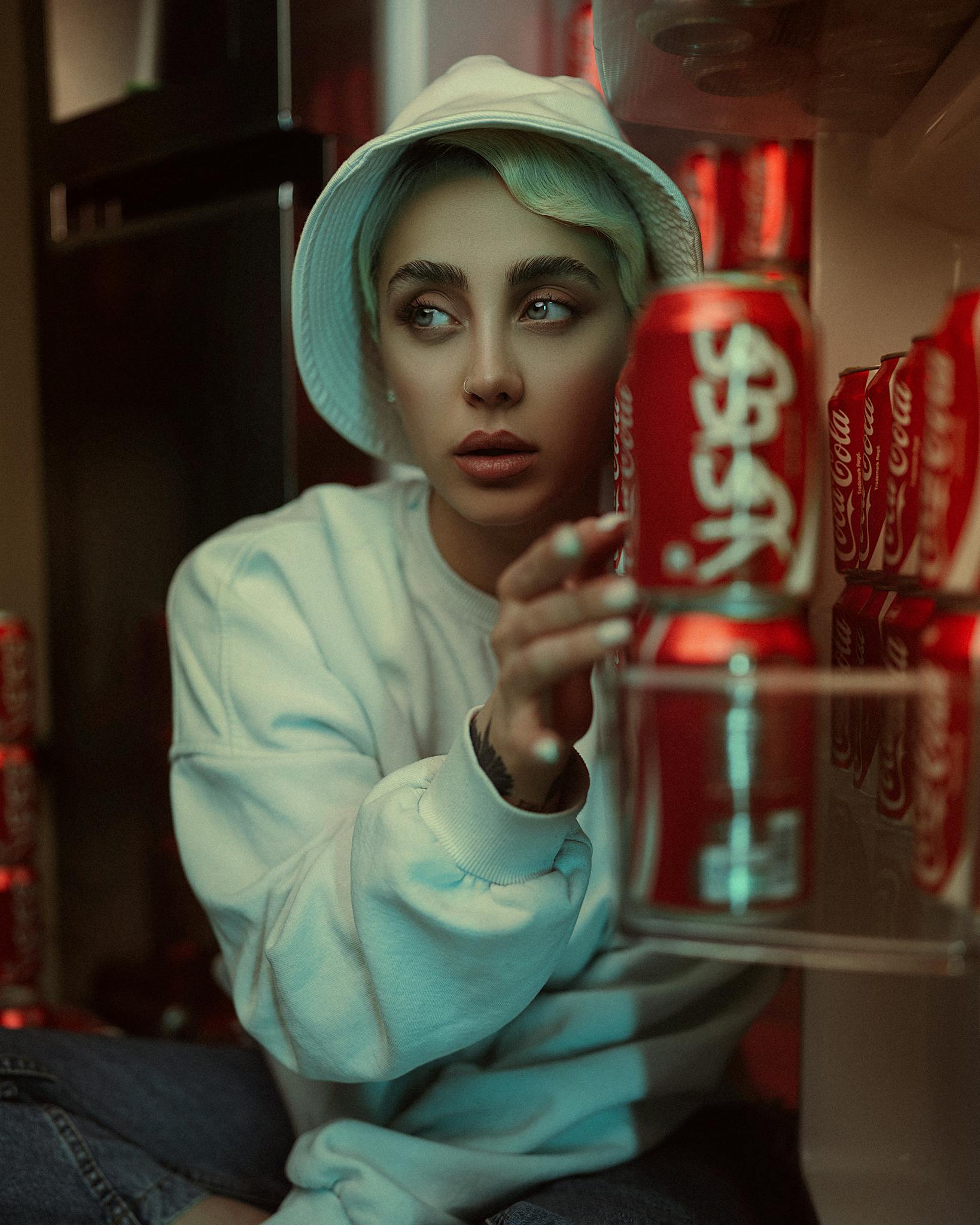 Stylish model with hat and pullover sitting near Coca-Cola cans indoors.